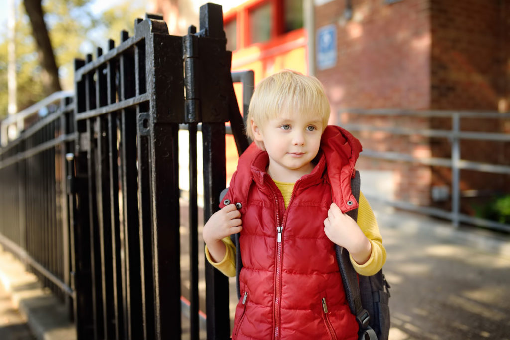 Young blonde-haired boy next to a school gate.